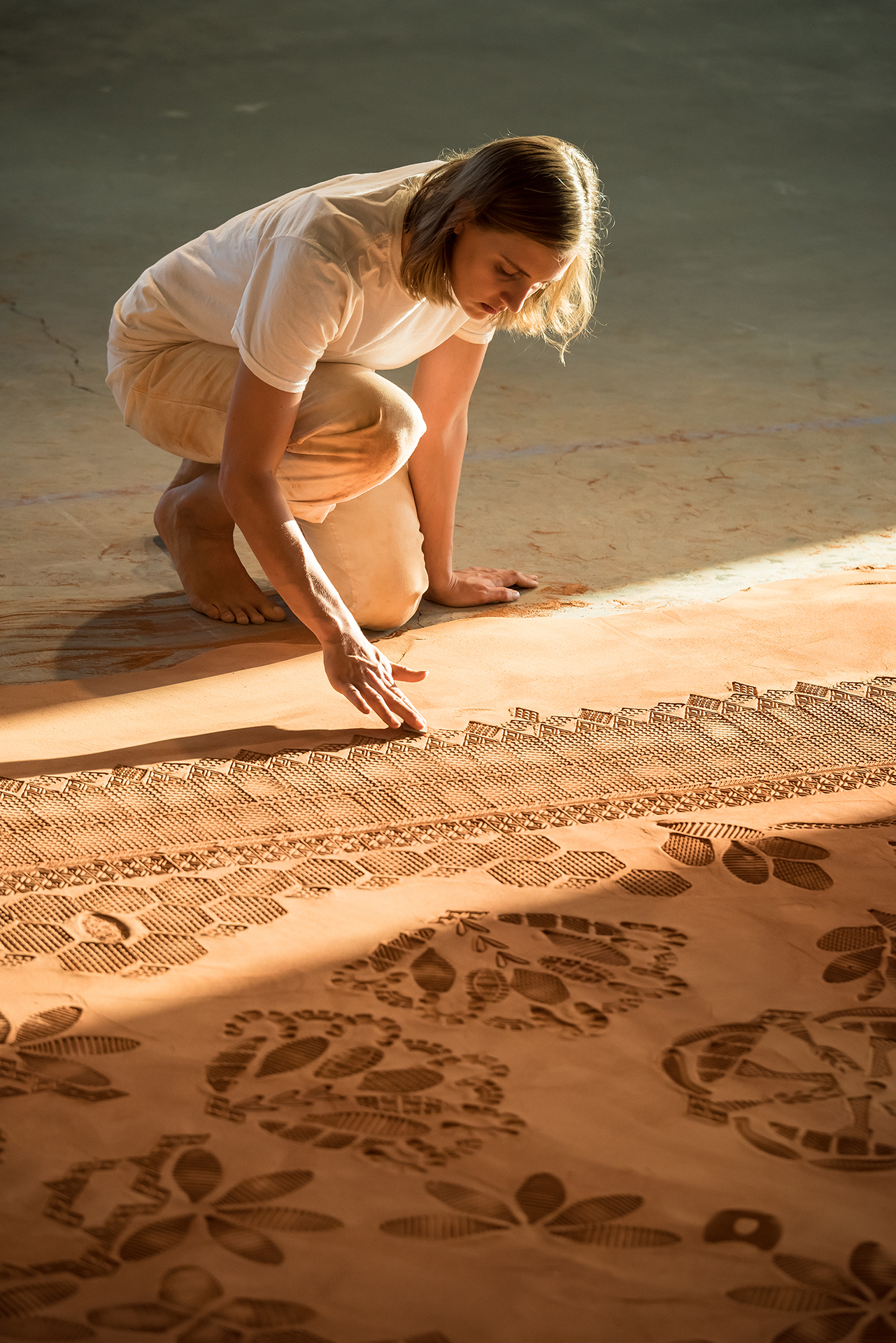 Photo of the artist working on a monumental, low relief sculpture mimicking an intricate patterned rug, made with modified shoe soles imprinting ground & sifted Oklahoma red dirt