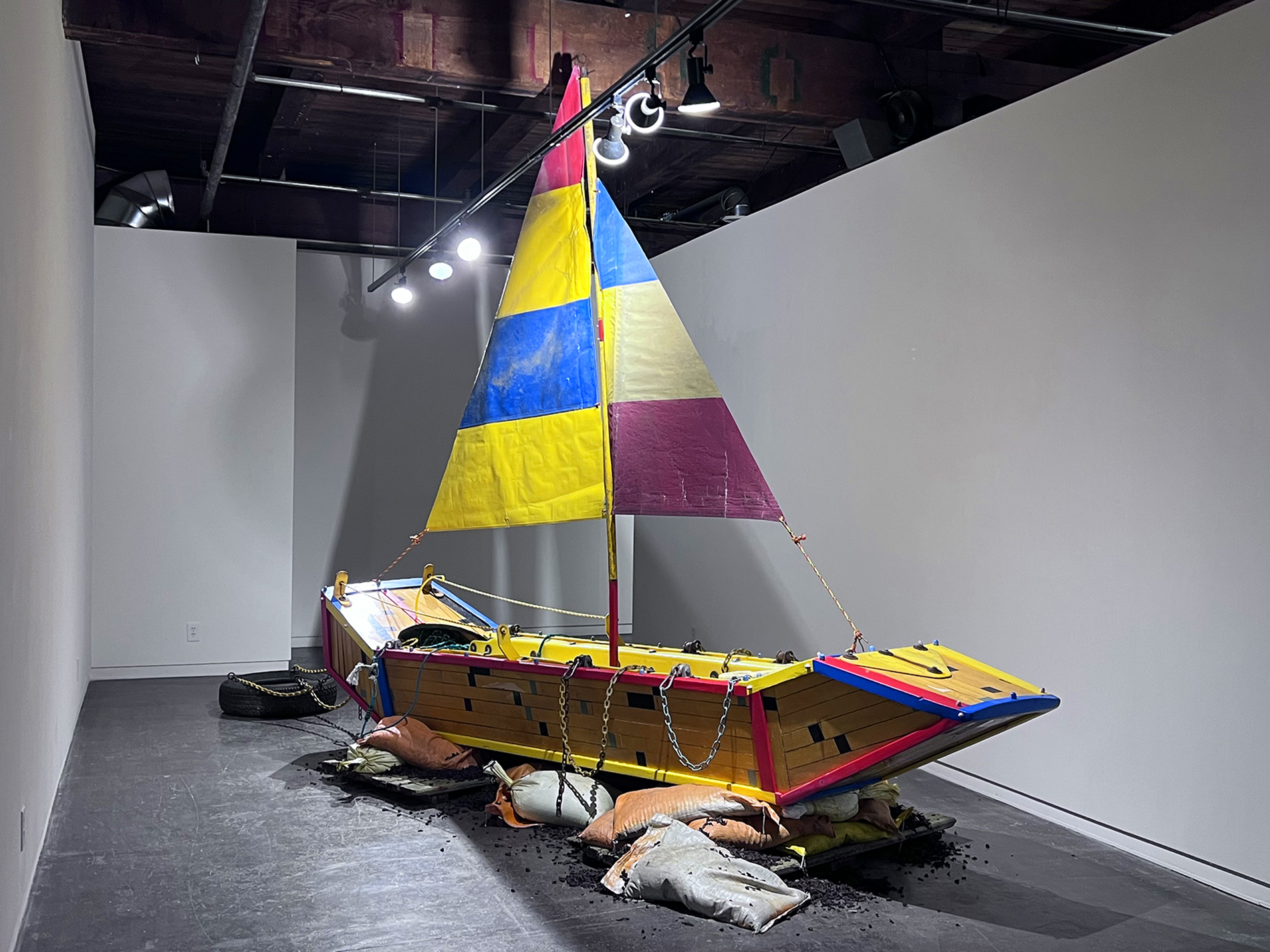 Aaron Coleman's Delicate and Filled with Dynamite, a handmade boat constructed from salvaged basketball court flooring is stranded on a sandbags filled with rubber mulch from turf football fields.