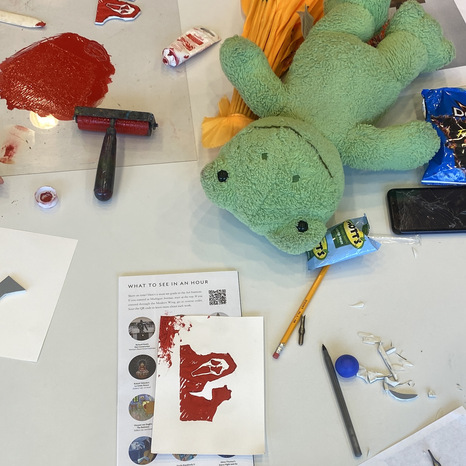 A stuffed frog lays on a work table surrounded by printmaking ink and brayer, and a small print with a Scream mask. Taken during a Teen Studio Workshop: Halloween Block Prints at Art Institute Chicago