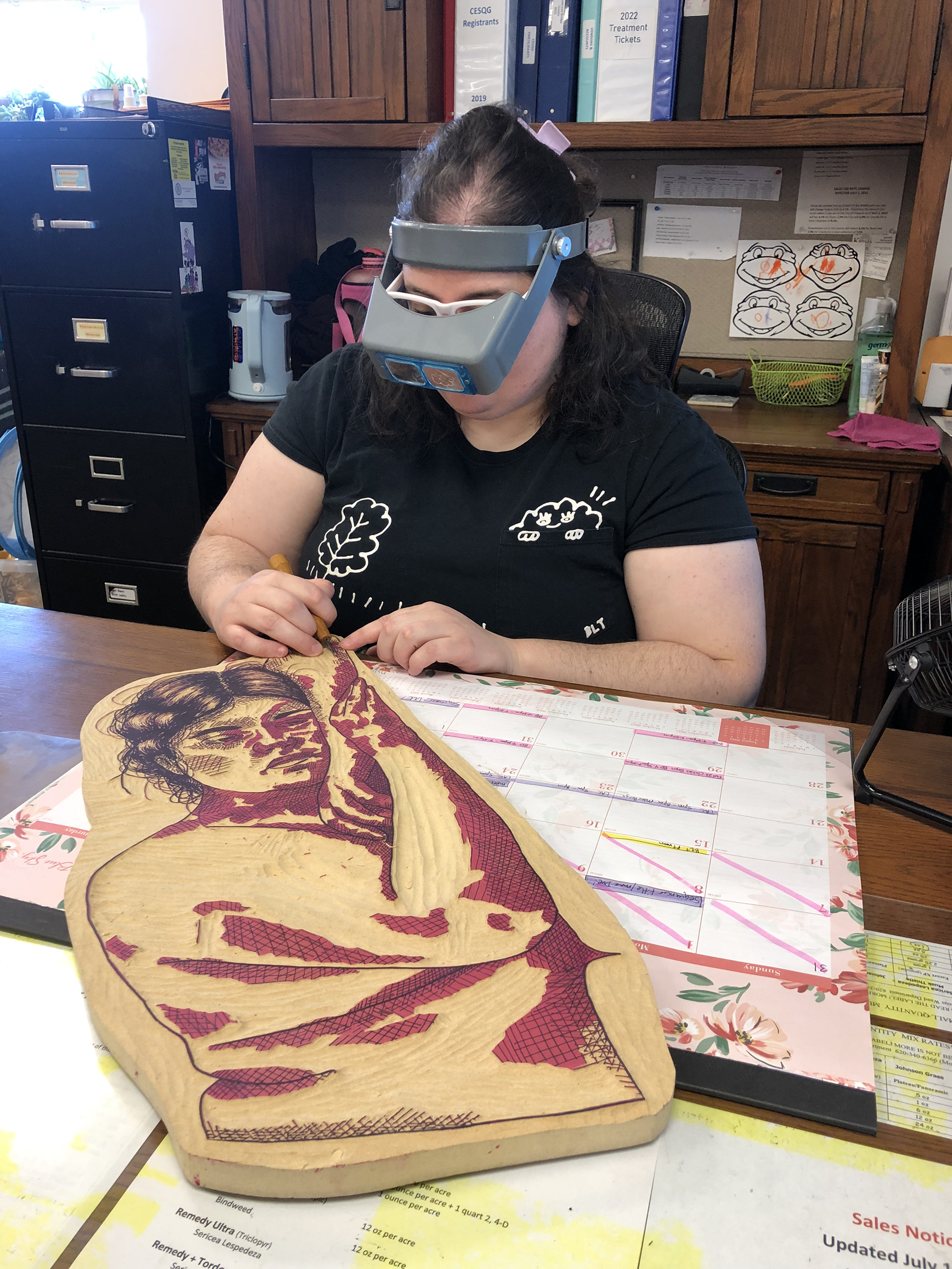 The artist wears a headband magnifier while carving a figure block.