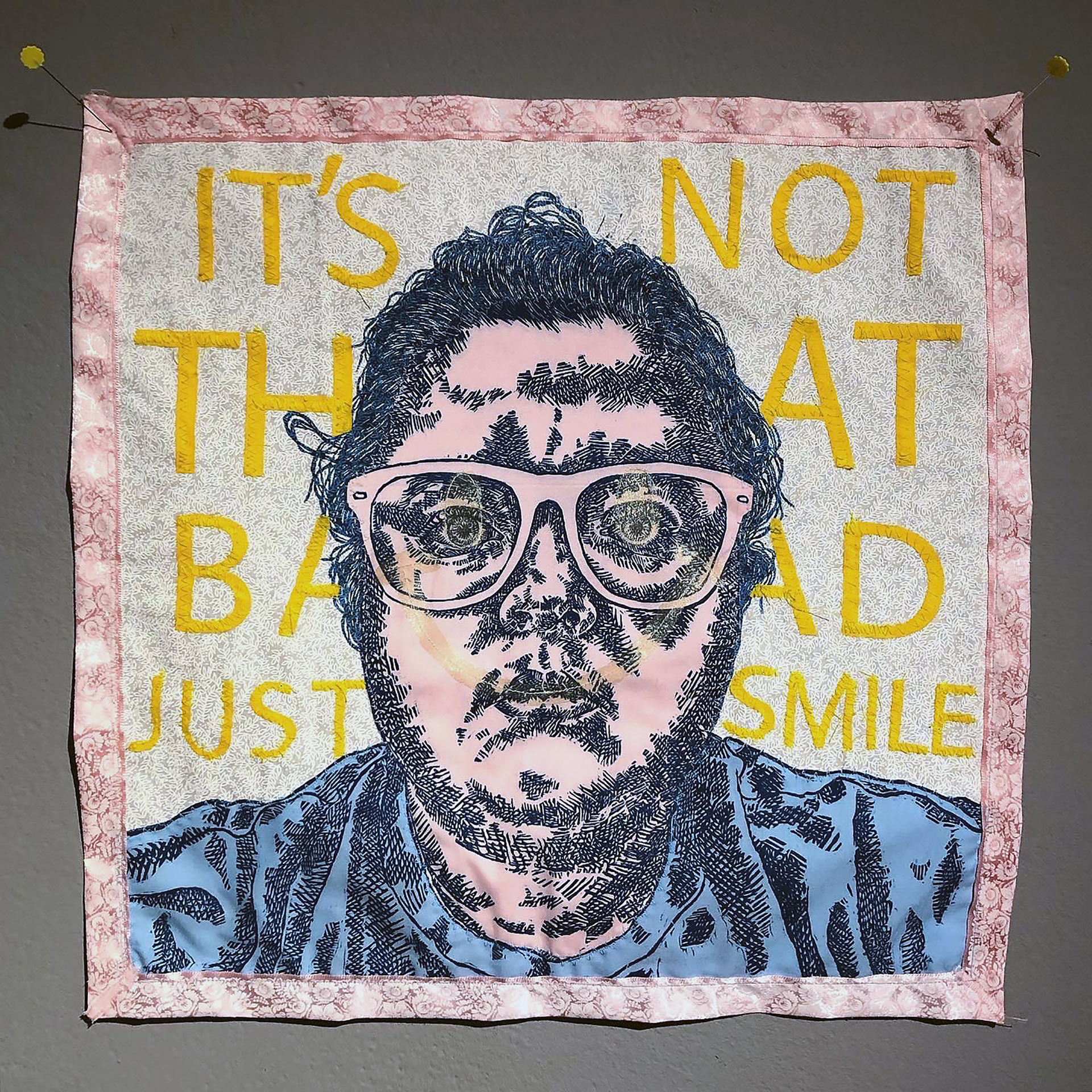 A relief printed portrait of the artist is printed on pink and blue fabric and appliquéd onto a quilt with the phrase "It's not that bad, just smile" stitched behind the figure. A subtle smiley face overlays the artist's face.