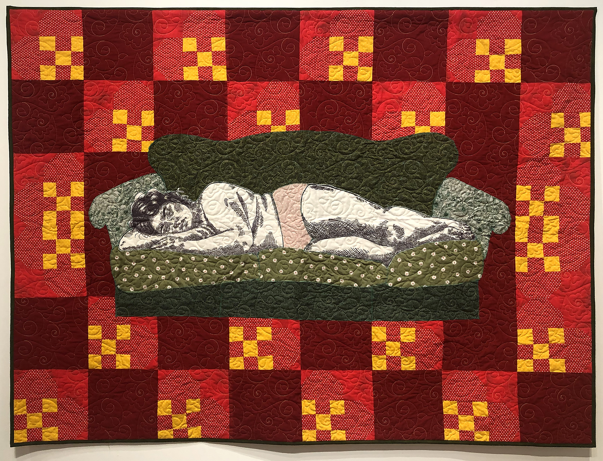 A sleeping figure lays upon a couch composed of quilted green fabrics. The sofa is stitched on top of a red and yellow quilt top.