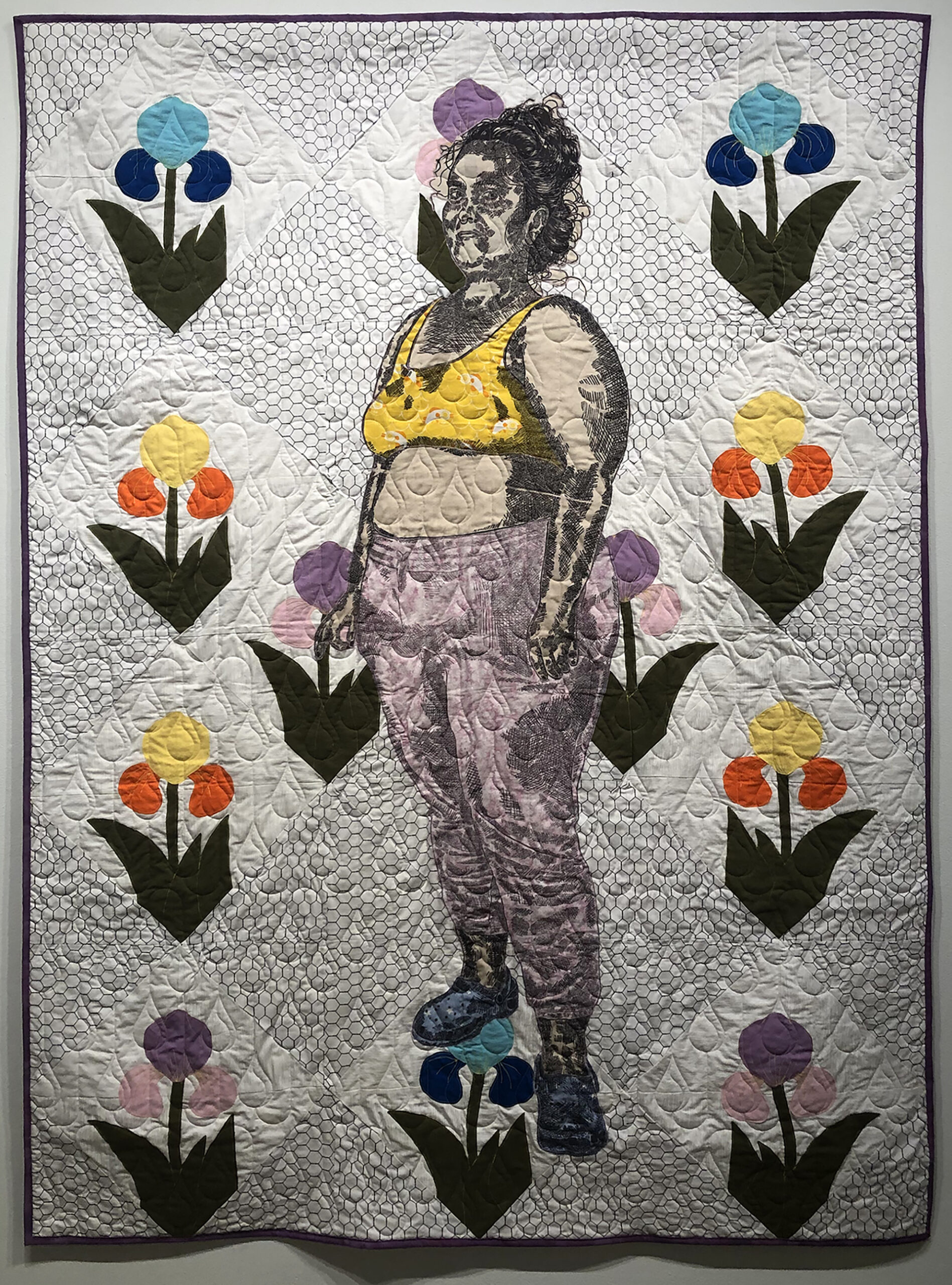 A standing figure printed on pink, beige, and yellow fabric, is stitched upon an elaborate floral quilt.