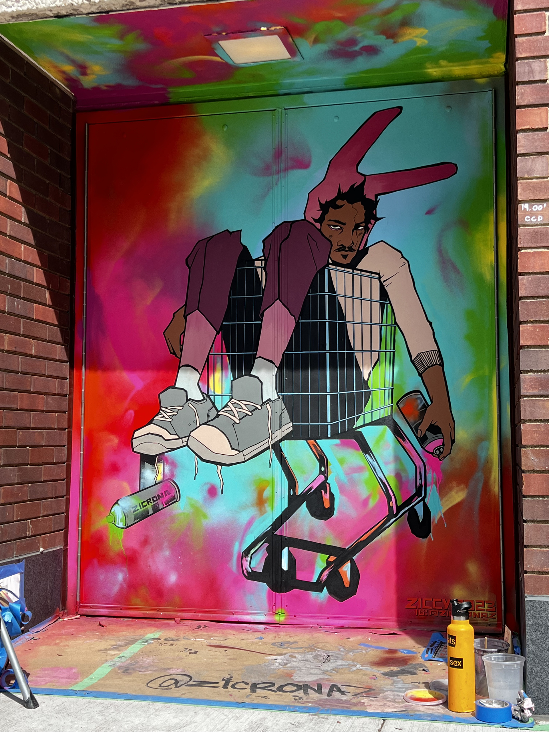 A mural on a doorway featuring tie-dye like spray paint and a figure wearing bunny ears laying in a shopping cart with spray paint cans.