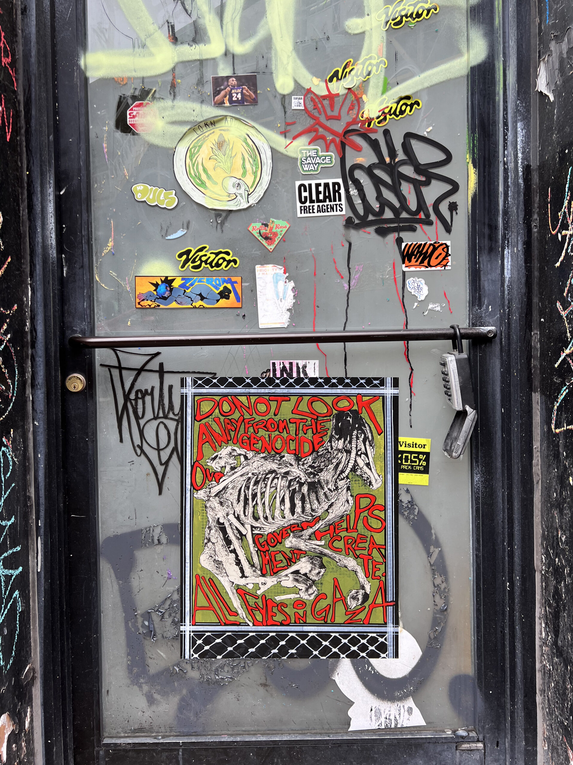 A paste up on a graffitied door of a dog skeleton surrounded by the stylized text "Do not look away from the genocide helps create. All eyes on Gaza"