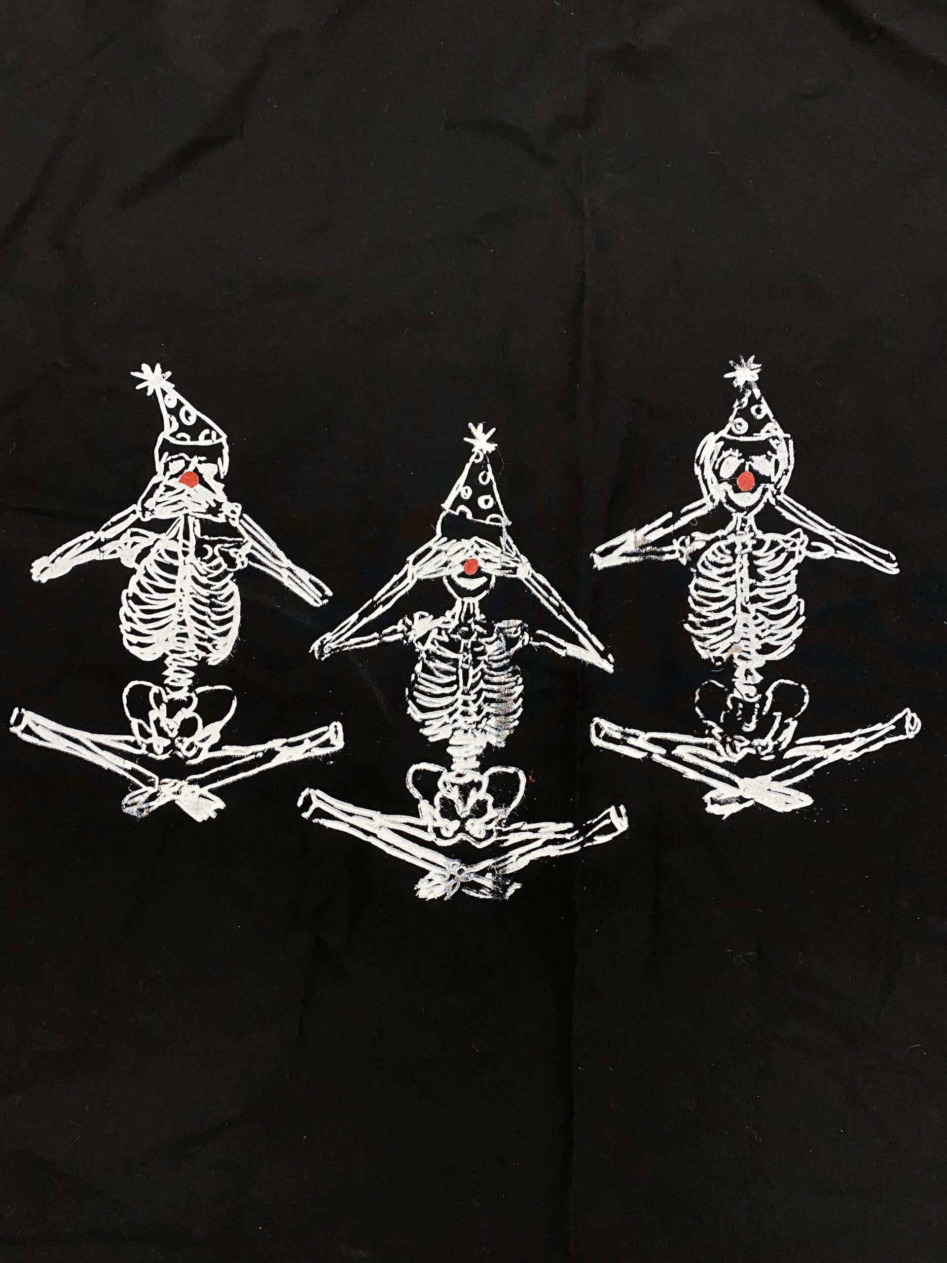 Three skeletons wearing party hats and clown noses are posed as Speak no evil, see no evil, hear no evil. Printed by a student at Marwen in white on black fabric