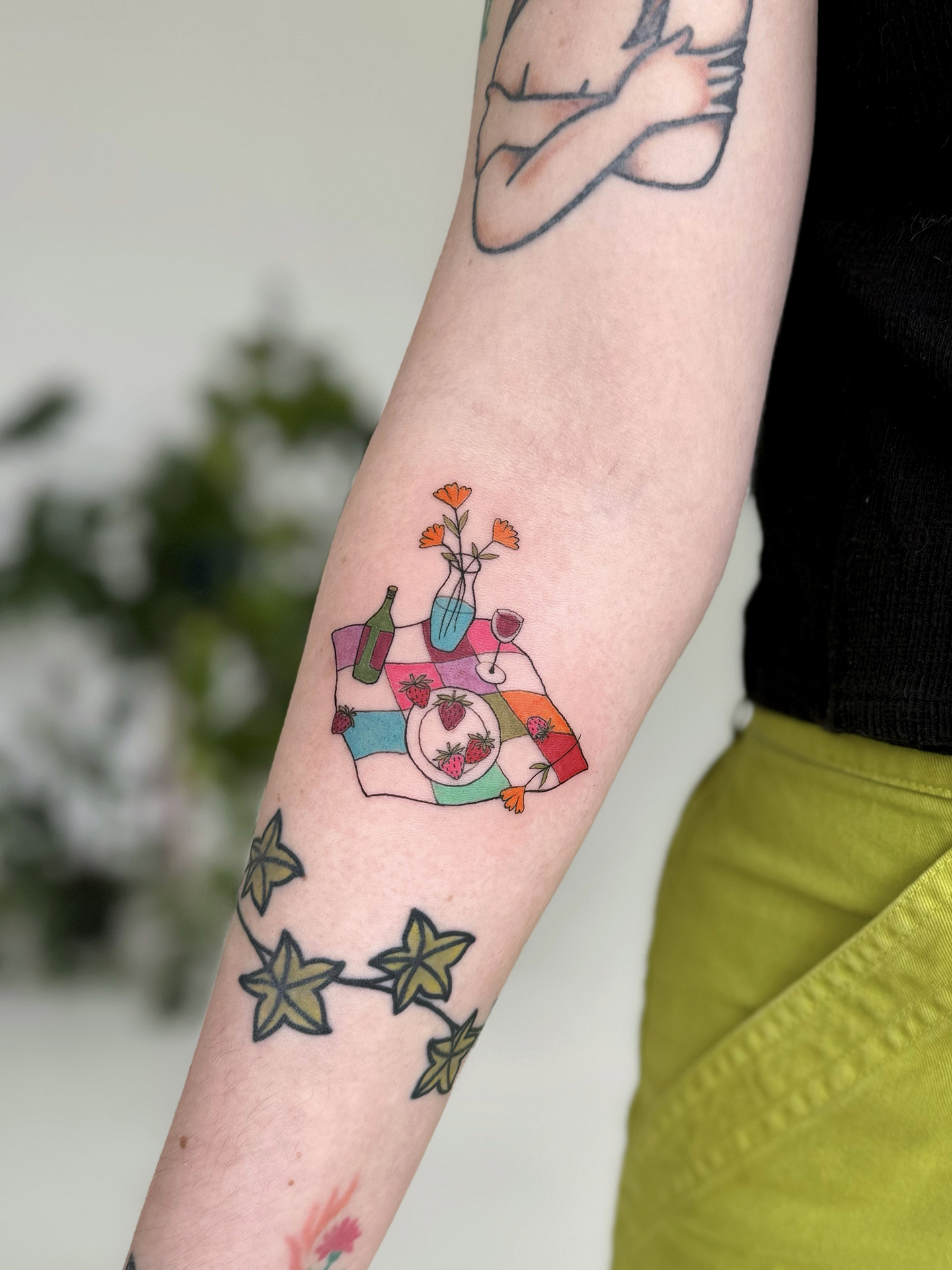 A colorful picnic blanket with strawberries, wine, and flowers is tattooed on a forearm.