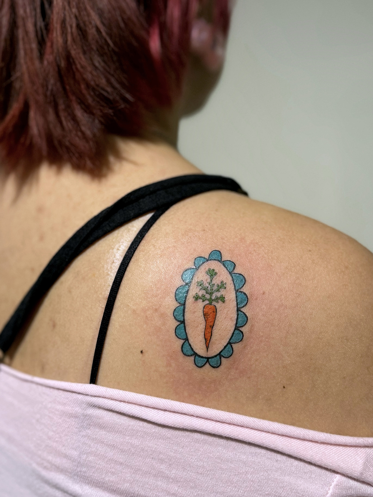 A single carrot, surrounded by a teal scalloped frame is tattooed on a shoulder.