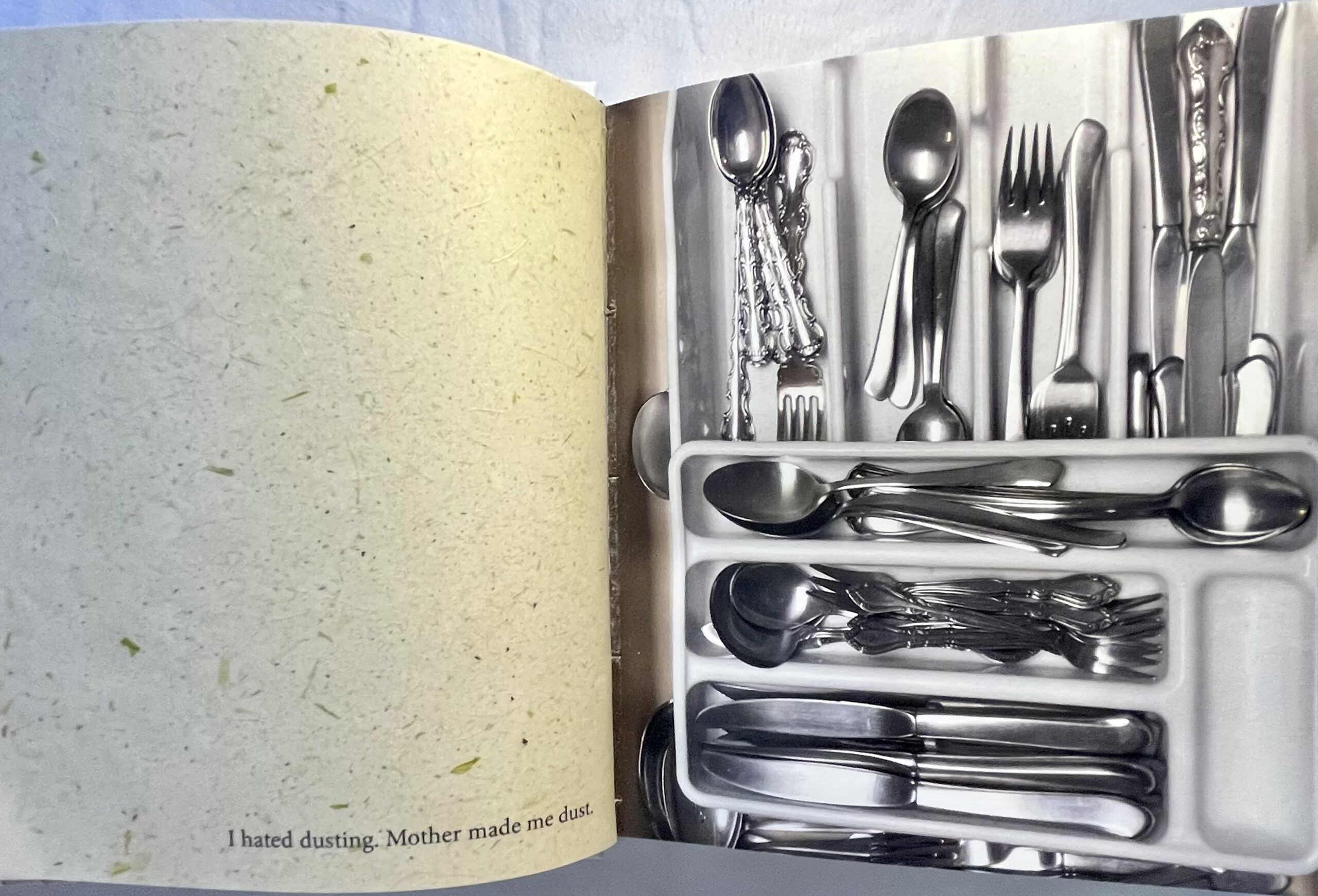 An open page of a book. On the right, a color photograph of a silverware drawer, and on the left, a page of tan transparent paper speckled with plant matter. On the bottom of the left-hand page, printed text reads “I hated dusting. Mother always made me dust.”