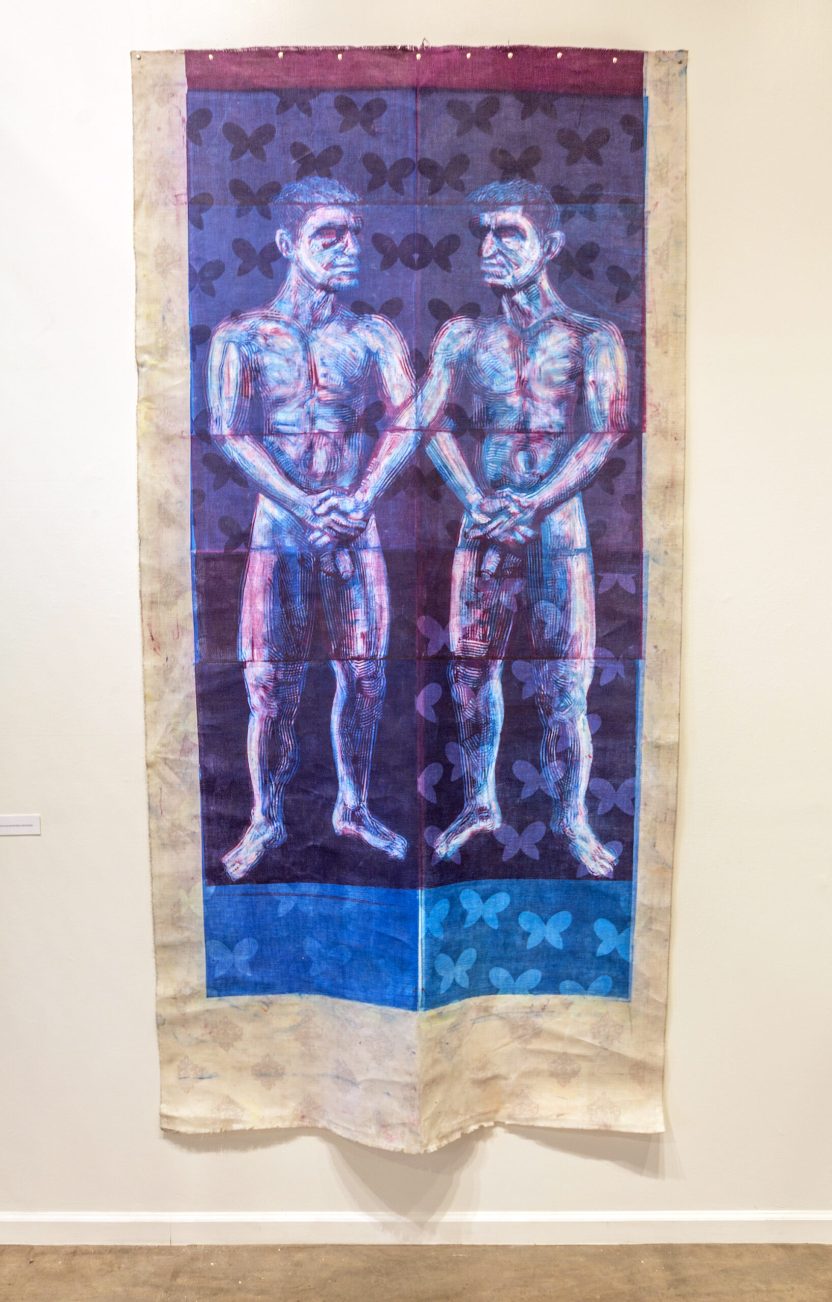 A pair of nude men, monoprinted in red and blue, hold hands and interlock arms. The fabric the figures are printed upon is covered in a pattern of butterflies, blockprinted with wax to create a resist.