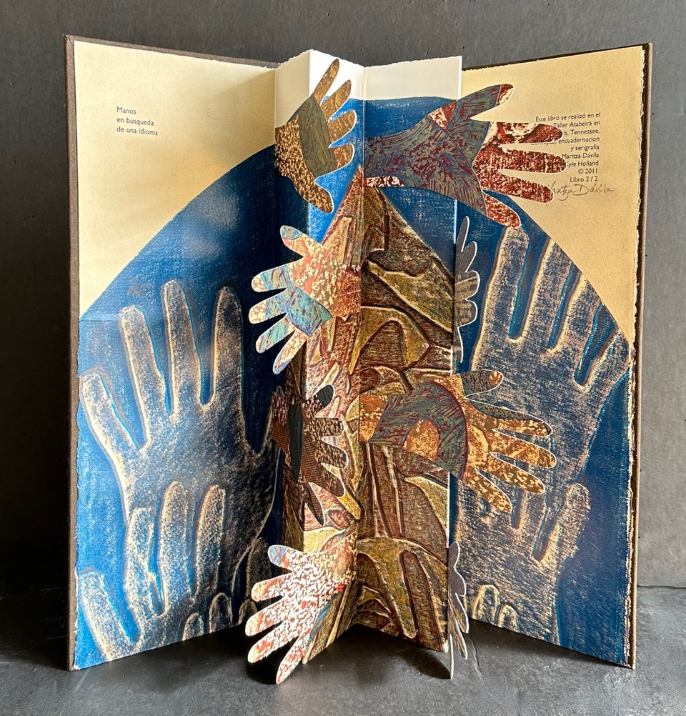 An accordion book with a pop-up center panel of hands outstretched and overlapping from the spine toward the edges of the book. On the primary pages text is above a half circle in blue filled with hands.