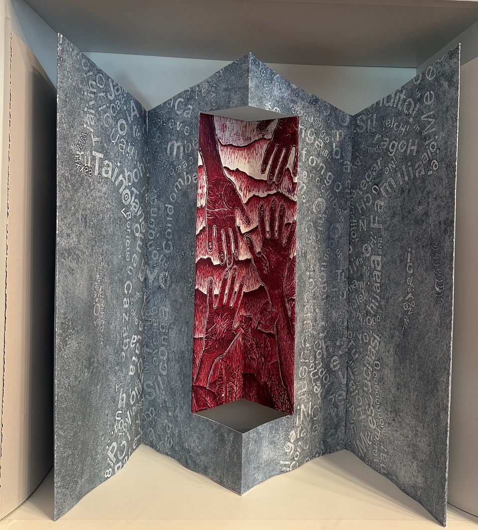 An accordion book with primary pages in gray text and a pop-up panel in burgundy with outstretched hands over strata of earth or rolling hills.