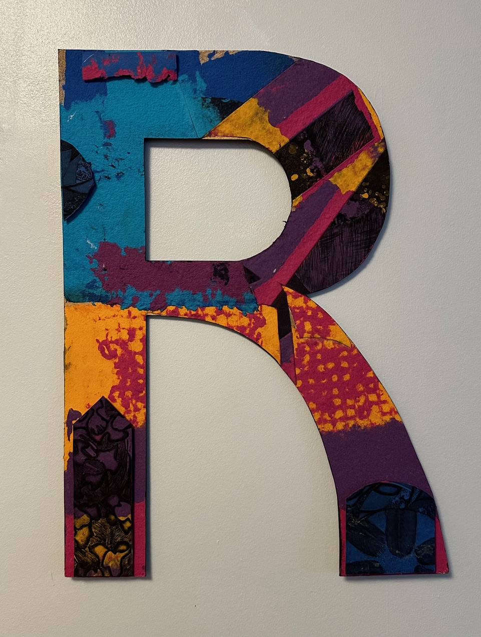 A large capital "R" is composed of bright colors and textures of handmade paper.