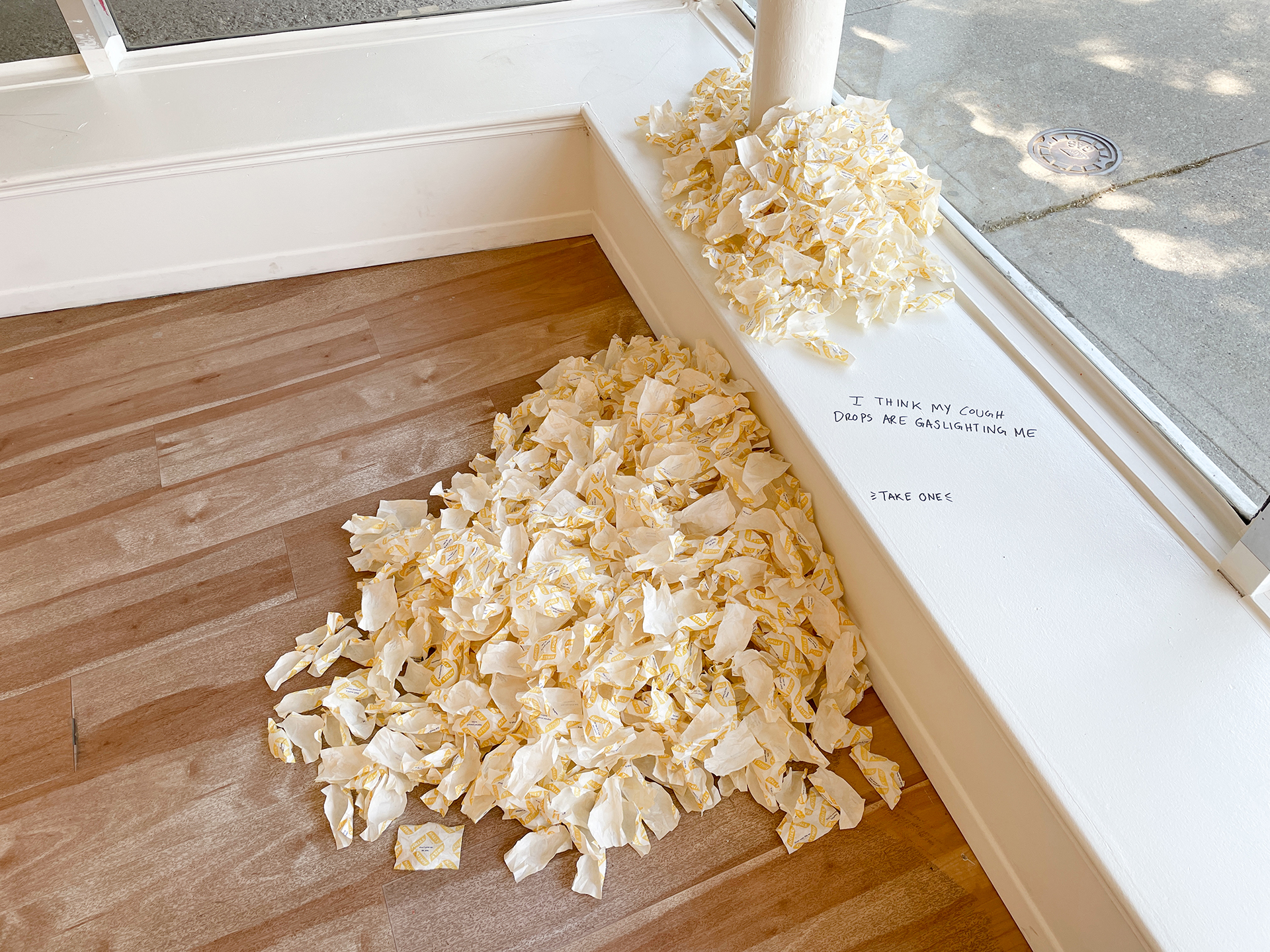 A pile of printed thin paper meant to mimic cough drop wrappers starts on a ledge and cascades onto the gallery floor. Text nearby encourages visitors to take one of the wrappers.