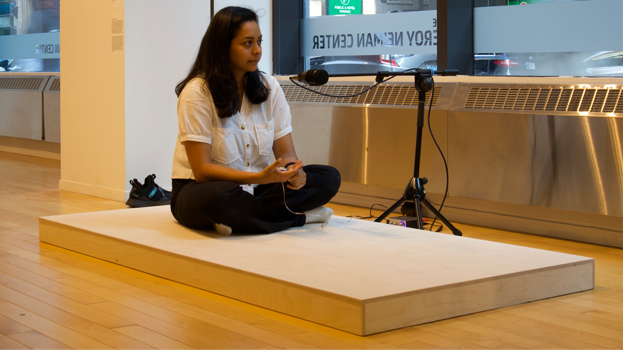 The artist sits on a platform with a microphone in front of her waiting for a viewer to join her.