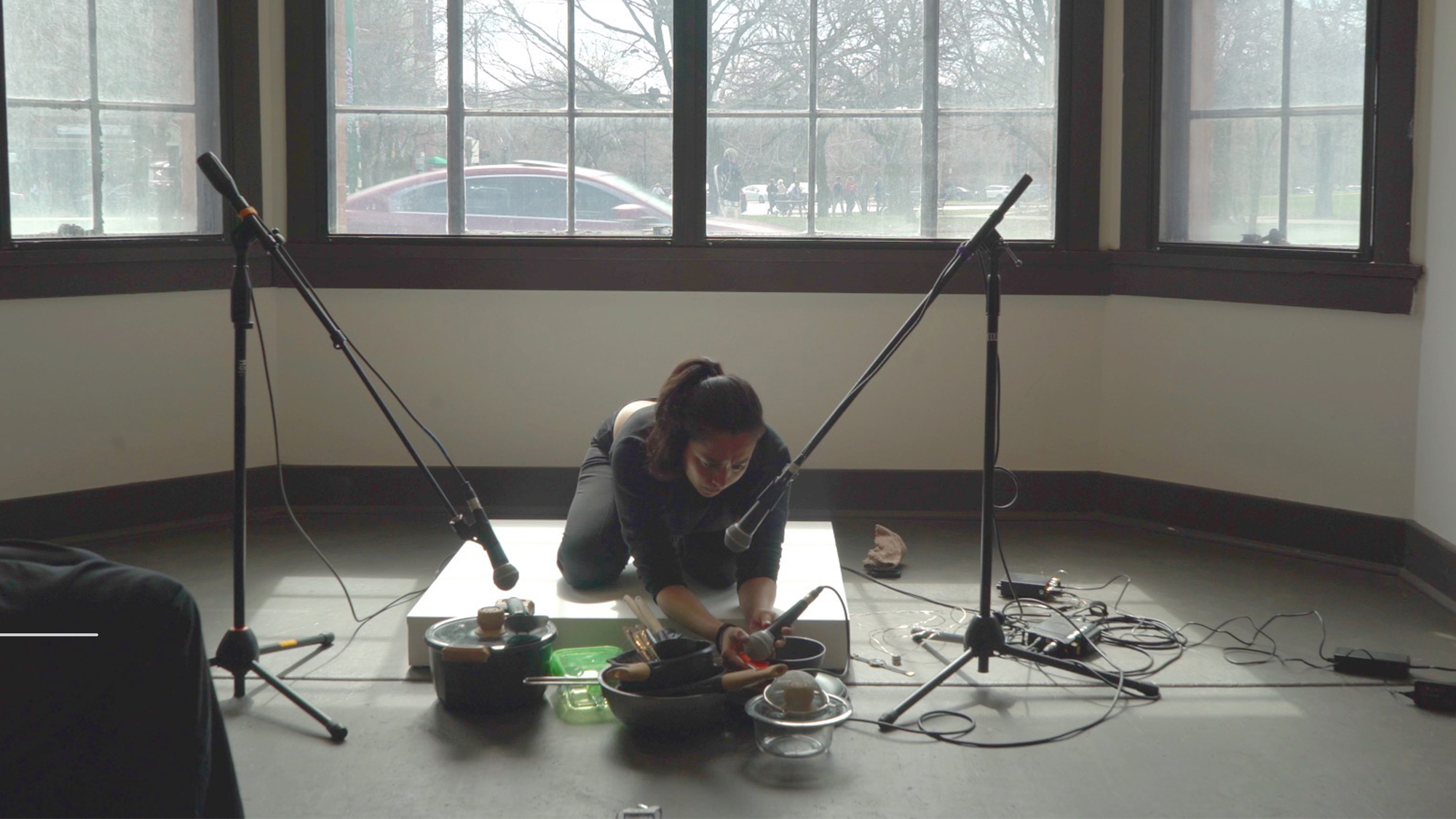 The artist sits on her knees, holding a mic in one hand and a utensil in the other, while performing.