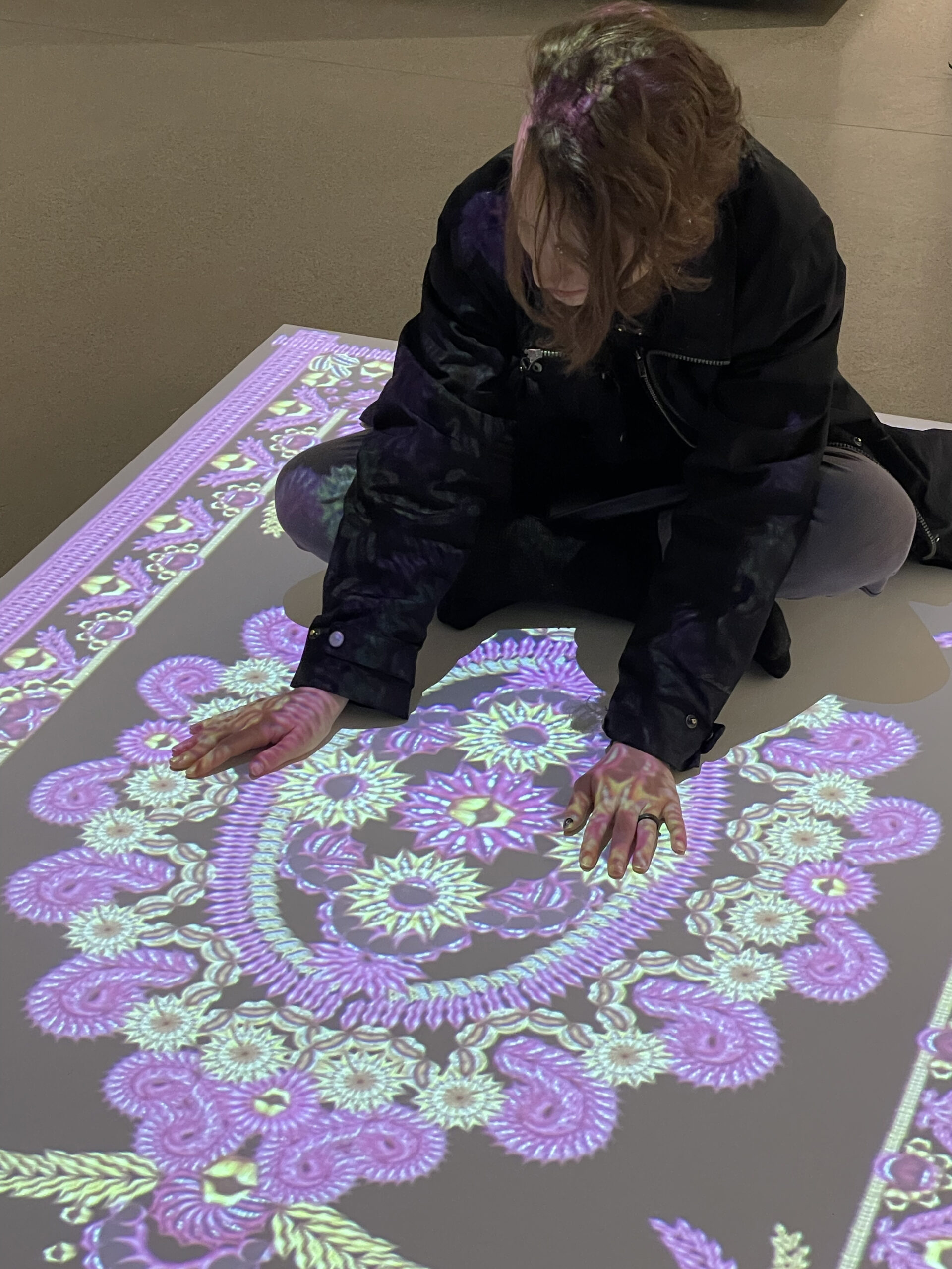 A person sits upon a platform with a two color overlaid with a dynamic projection featuring moving lip motifs in a rug-like pattern, with bright yellow and pink hues. The projection covers the hands and arms of the viewer.