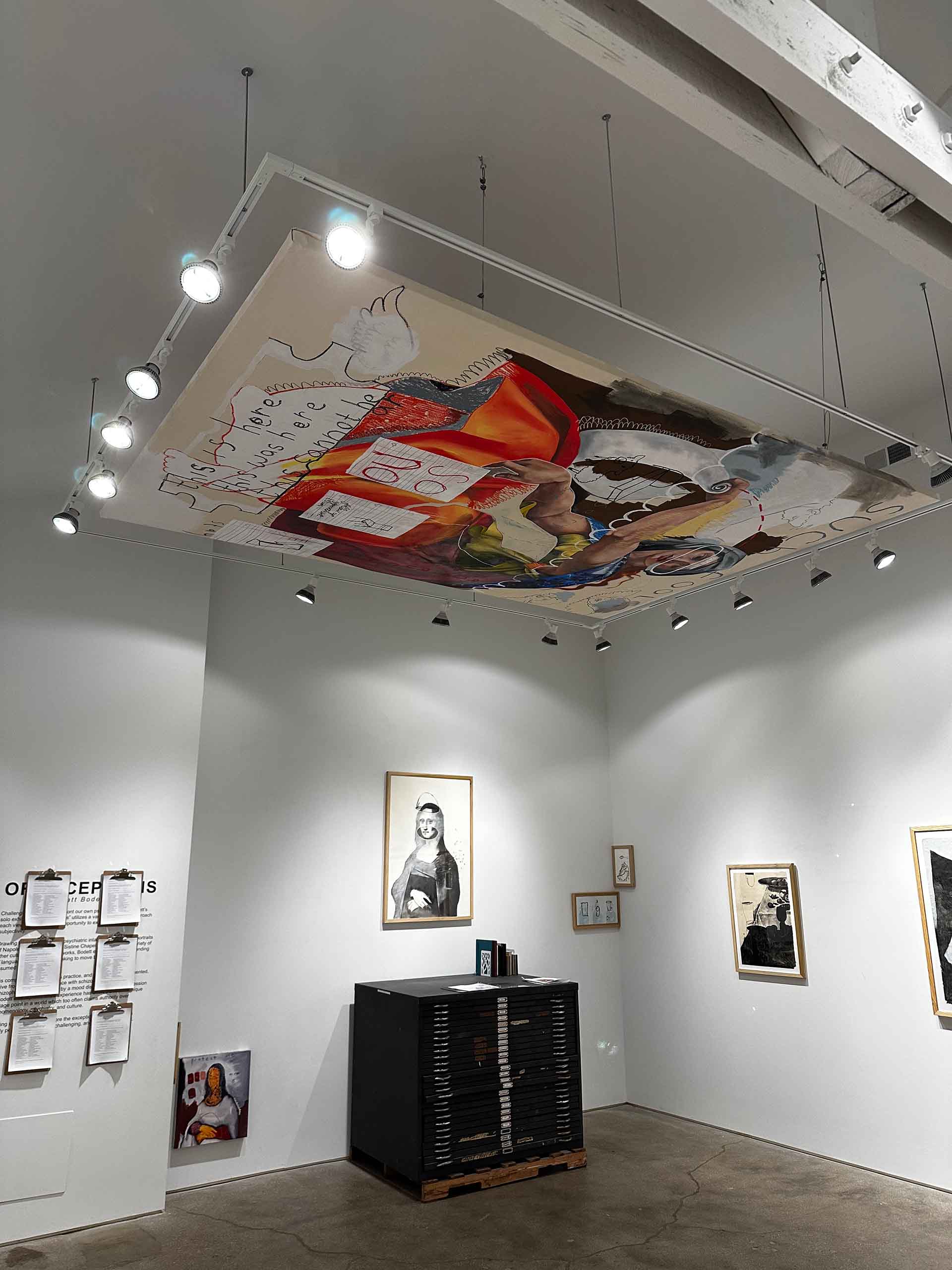 An installation image of the “Such Fools” painting. This is being hung from a ceiling, contained within a rectangle of track lighting. There is artwork hung at varying heights along the walls and a large flat file against one wall.