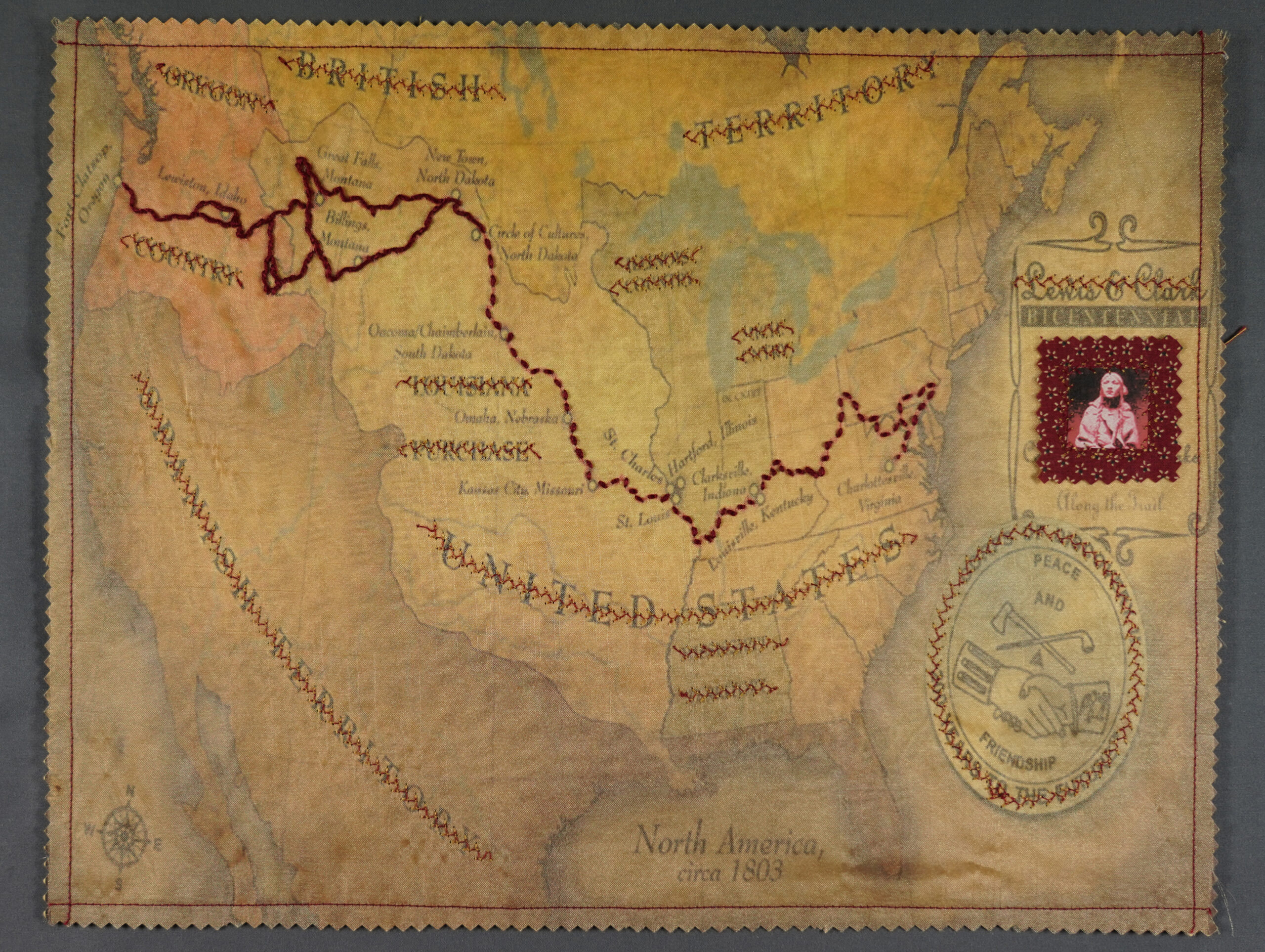 A map of North America from 1803 with the route of the Lewis & Clark expedition stitched in red thread through the fabric. The portion of the route that Sacajawea led is in bolder stitches. Colonial place names and Lewis & Clark have been stitched over as well, leaving an emblem that says "Peace and Friendship". A small image of Sacajawea is stitched into an inset claiming the bicentennial of the expedition as her own.