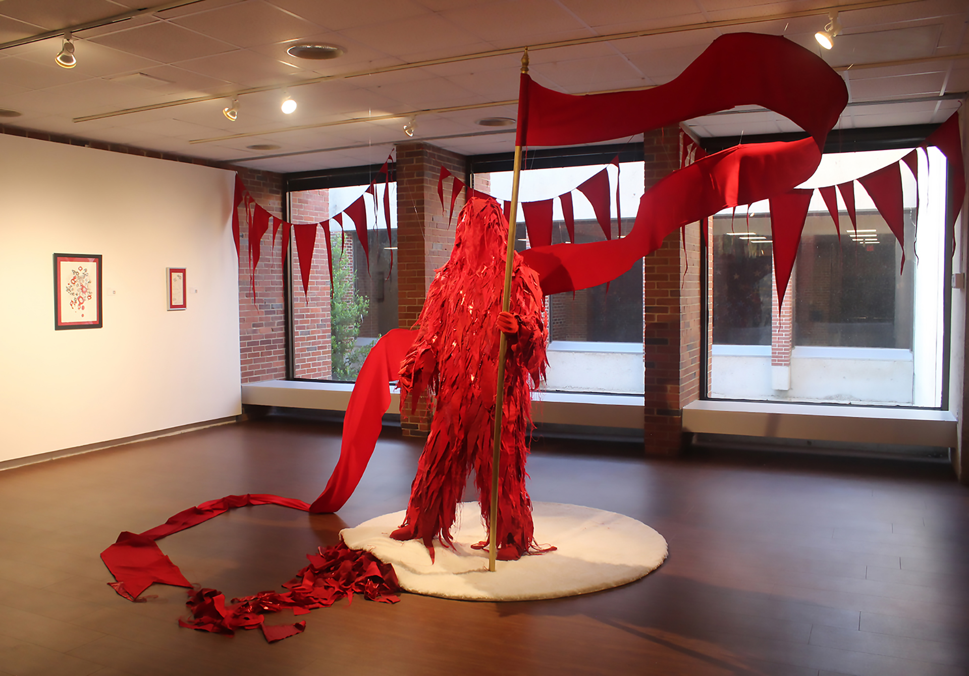 A sculpture of a standing figure covered with layers of red fabric hols a long red flag that piles on the floor.