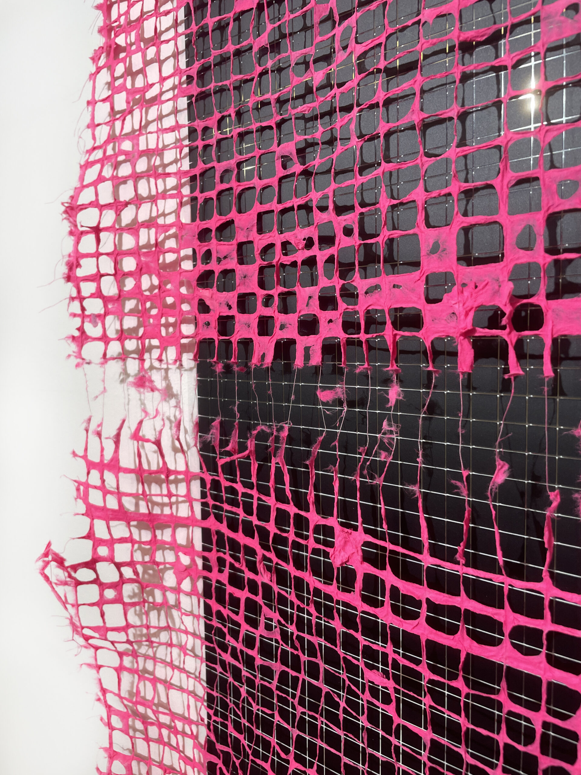 Closeup of a hot pink handmade paper grid handing in front of a vinyl grid comprised of many small black squares.