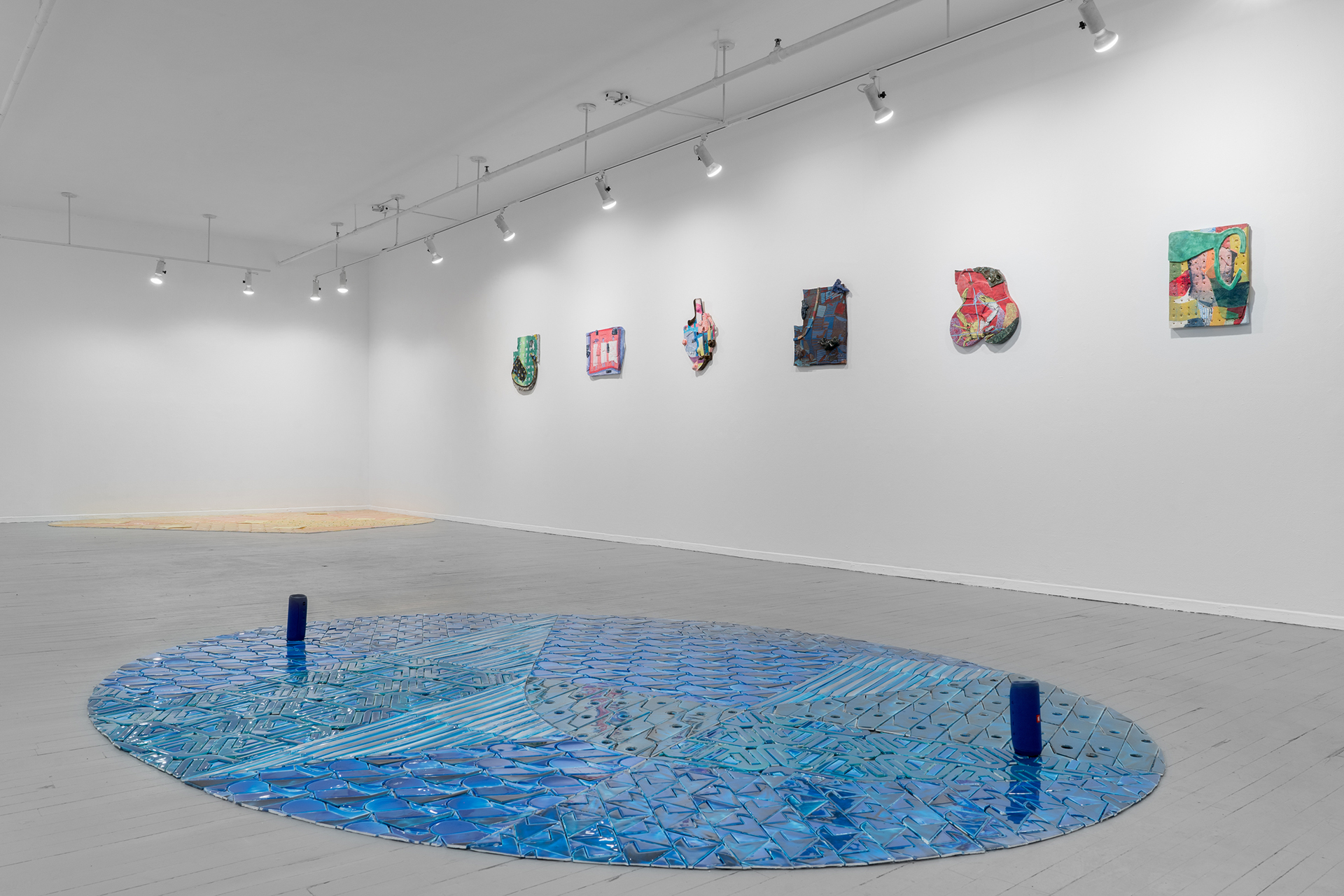 Installation view of Luedtke's exhibition with a blue oval on the floor evoking a pool and six shaped collaged works hanging on the wall.