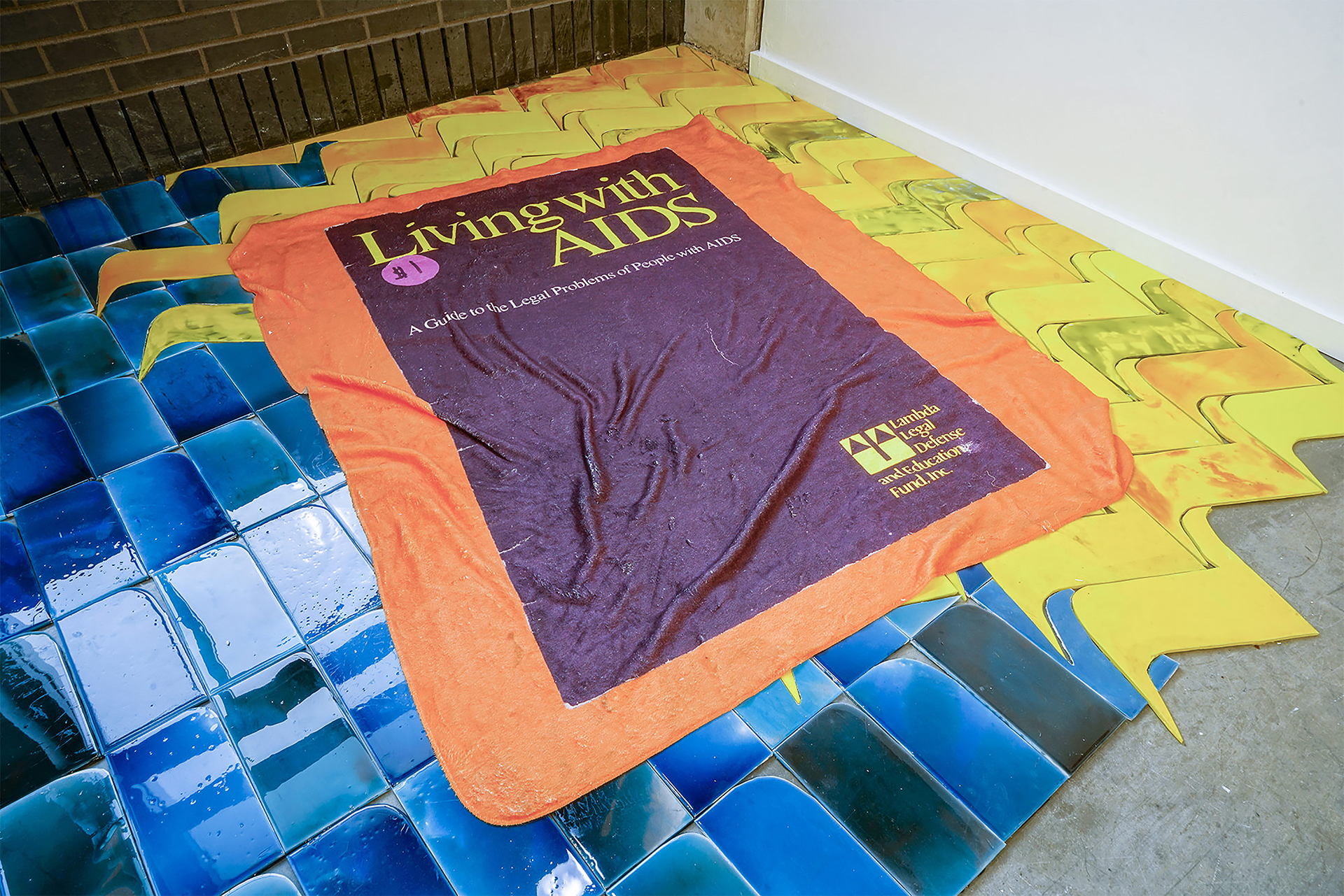 A blanket is printed to look like an enormous version of a guide called "living with AIDS", including a sticker that prices the book at $1. The blanket is laid out over a faux tile installation in blues in a sky-like grid, and yellows that create an abstracted sun.