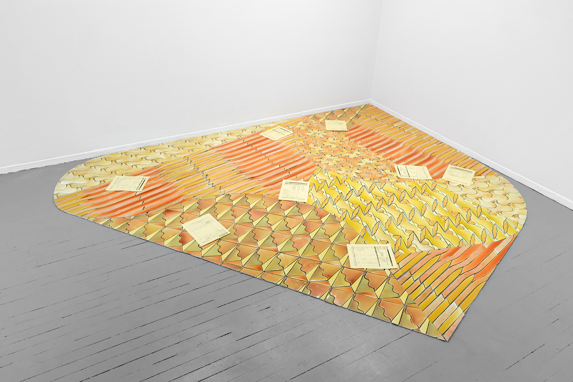 A large, rounded diamond floor installation with tile-like components in yellows and oranges. Scattered atop the tiles are copies of the artists' blood and urine analysis.