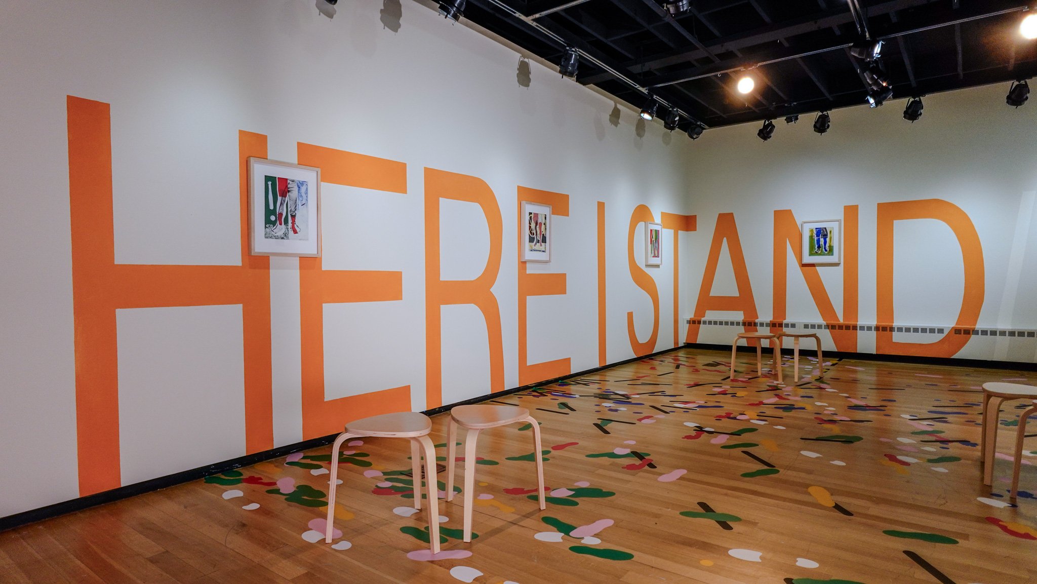 Four framed prints of legs at different stages of rehabilitation hang atop a 6’2” text mural stating “Here I Stand.” Six wooden stools are dispersed throughout the space on a wooden floor with a vinyl floor drawing depicting the footprints and imprints of different walking assists.