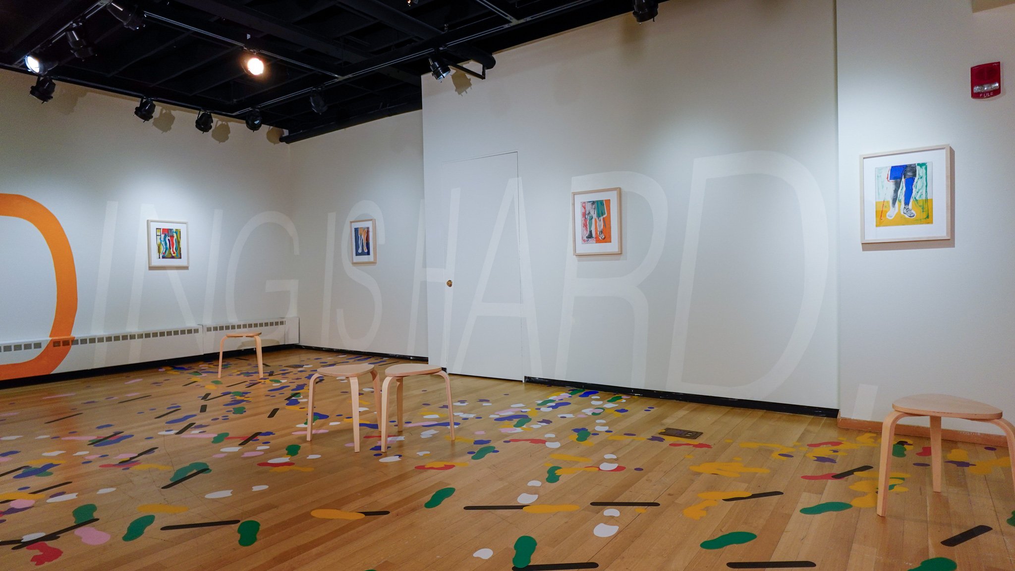 Alt Text: Four framed prints of legs at different stages of rehabilitation hang atop a 6’2” text mural stating “Standing Is Hard.” Five wooden stools are dispersed throughout the space on a wooden floor with a vinyl floor drawing depicting the footprints and imprints of different walking assists.