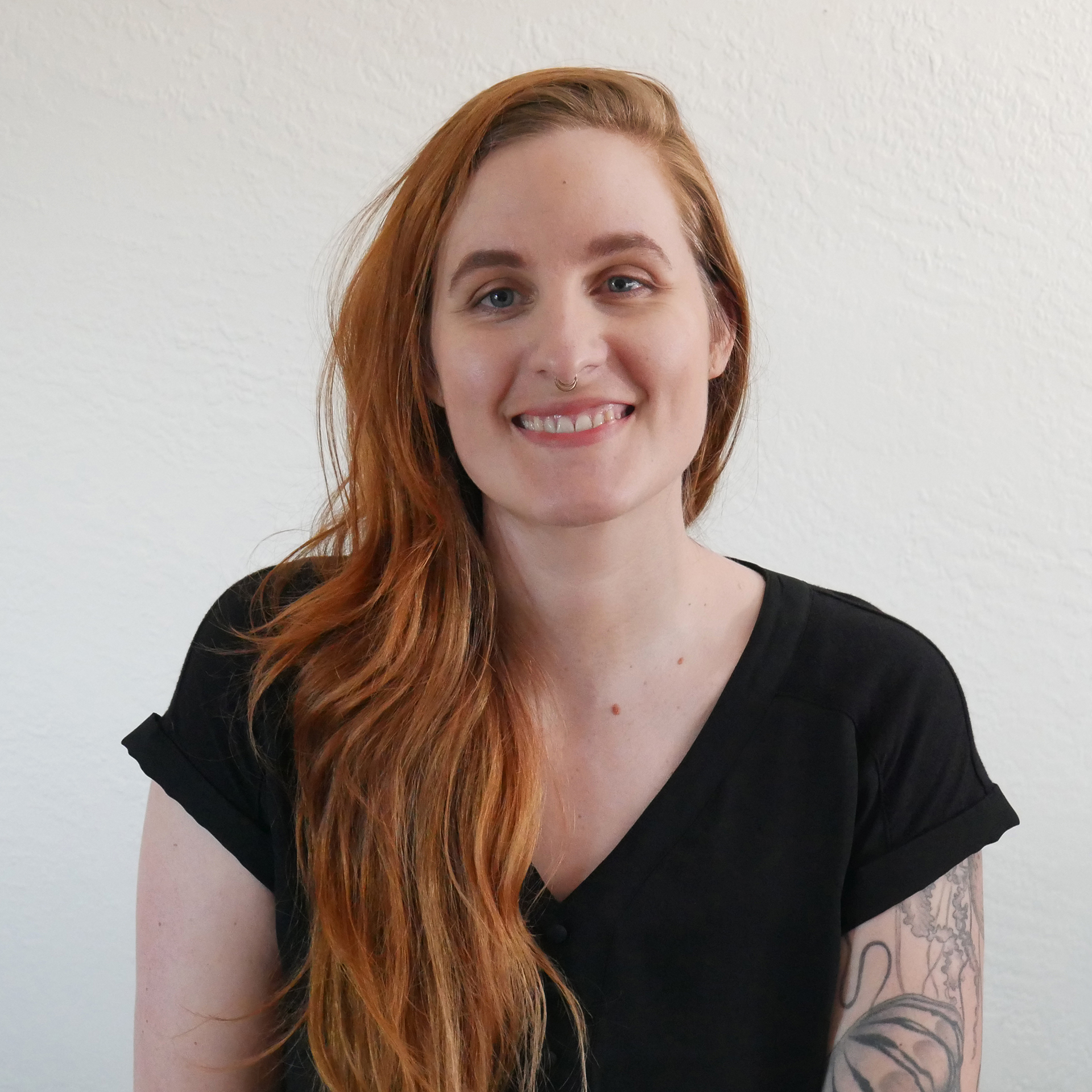 A portrait of Eliza, a white person with long, wavy, red hair pulled to one side wearing a black T-shirt and smiling.