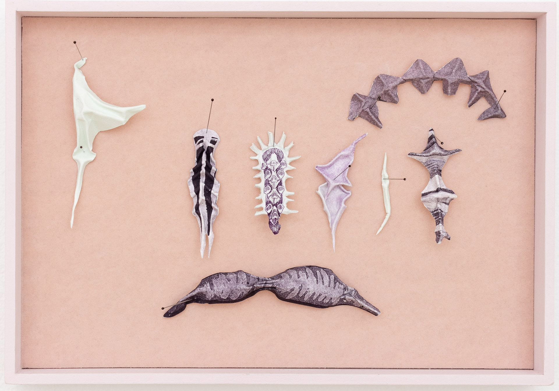 Handmade objects evoking talismans or alien insect specimens sit in a shadow box frame.