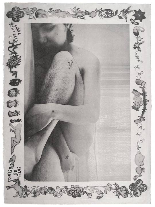 The artist is curled up nude on the floor surrounded by a frame of icons and doodles