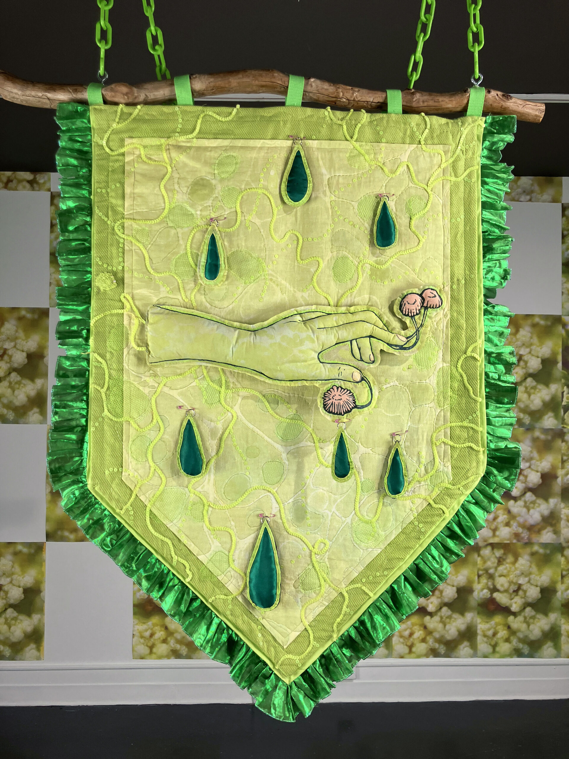 A banner in bright greens, with a hand w/mushrooms growing from the fingers and raindrops surrounding it, hangs from a found stick and green chain. A wallpaper installation is behind.