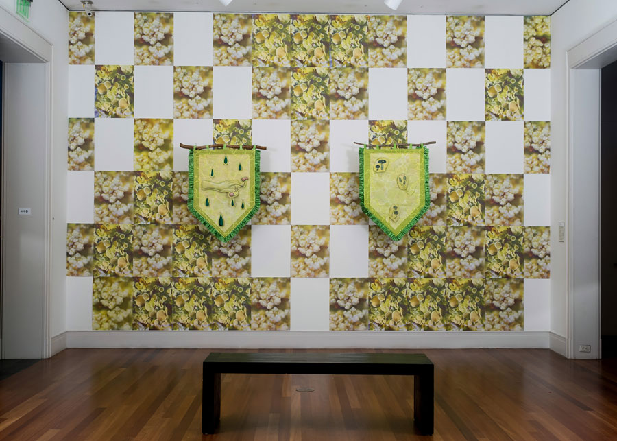 Green banners on found branches are mounted in front of a wall paper installation of foliage with panels of the the paper missing to create an irregular white grid