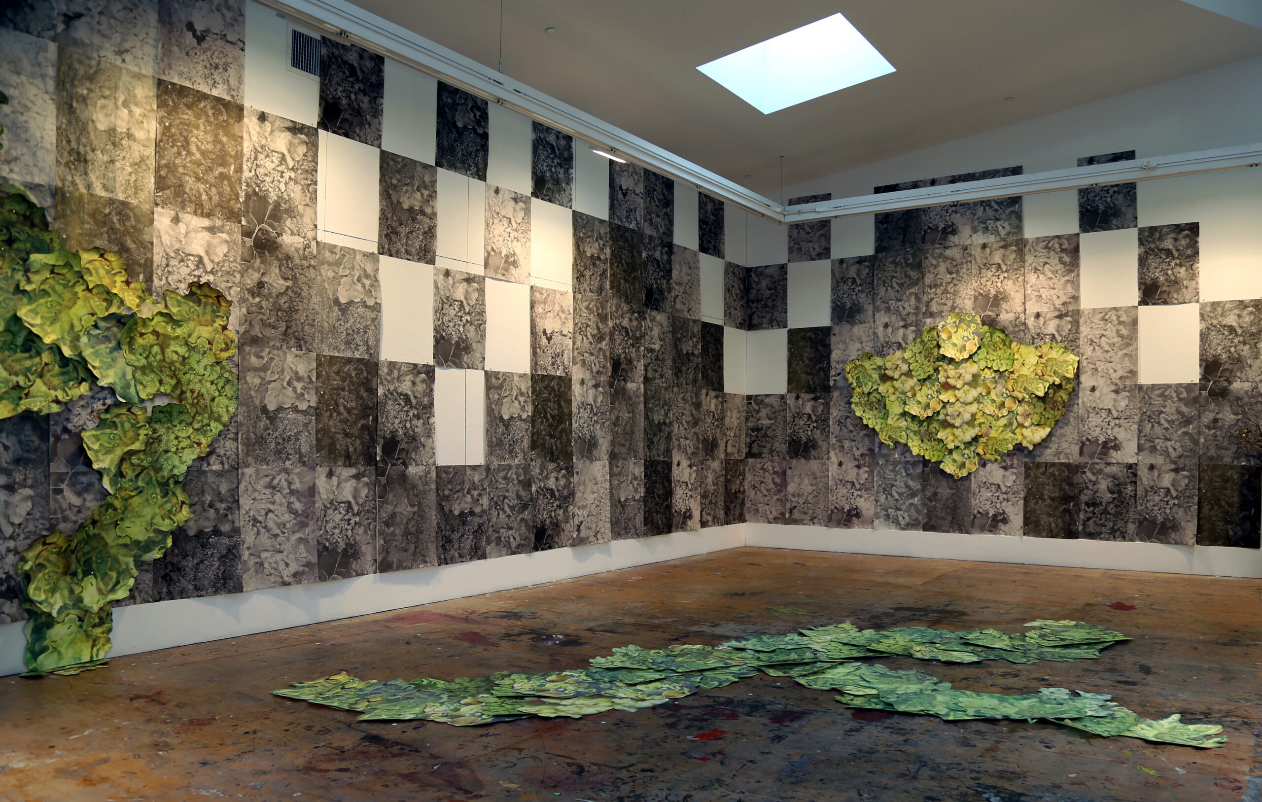 Enormous green printed shaped sculptures evoke moss sitting on dark wallpaper of mold with panels missing to create an irregular white grid. Other "moss" sits on the floor as well.